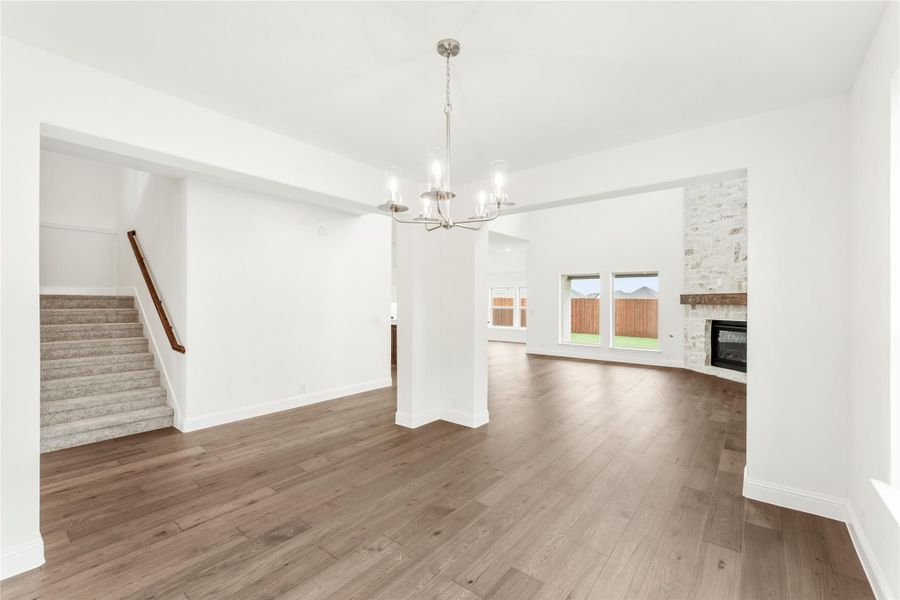 Open living and dining area with hardwood floors, stone fireplace, chandelier, and staircase nearby.