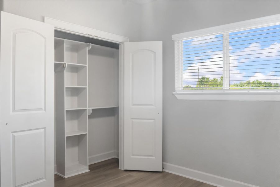 A bright room with light gray walls and wood flooring features a window with blinds offering a view of greenery. It includes a built-in closet with shelves and hanging space behind white double doors.