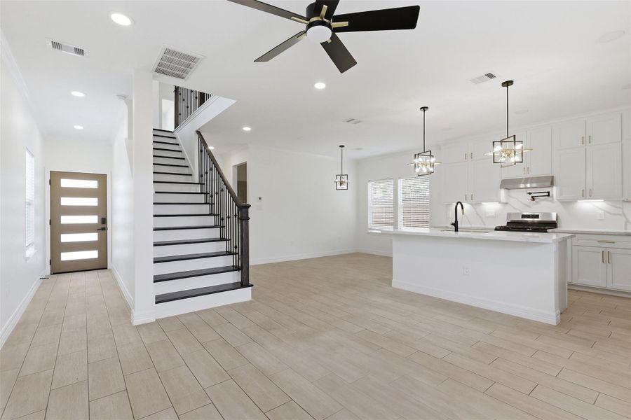 Bright and open first floor layout showcasing elegant staircase with black iron railings, ceiling fan, and kitchen island beyond.
