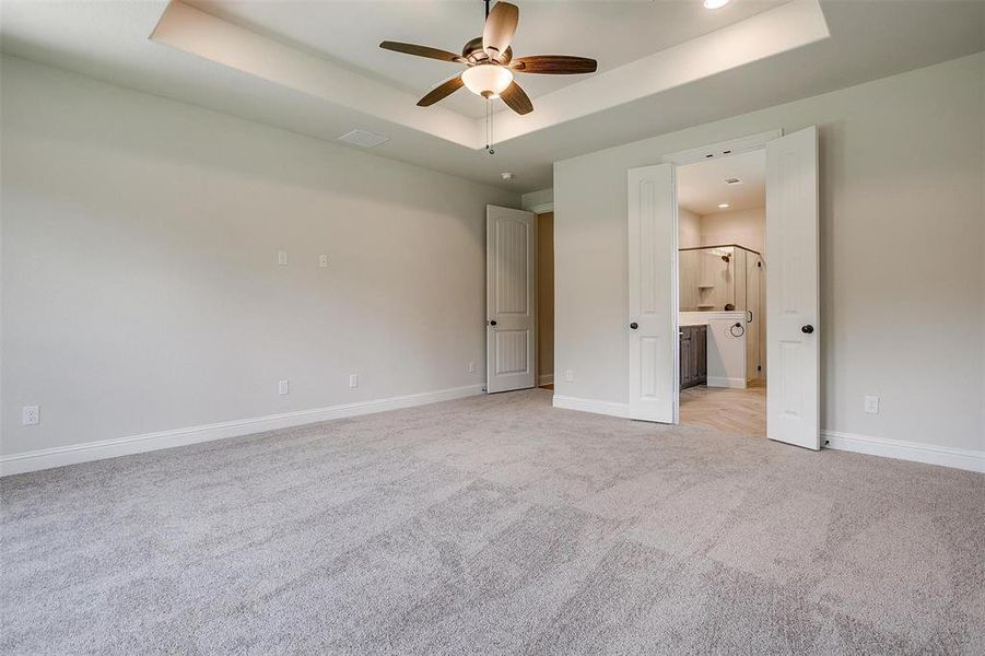 Unfurnished bedroom with light colored carpet, ensuite bathroom, ceiling fan, and a raised ceiling