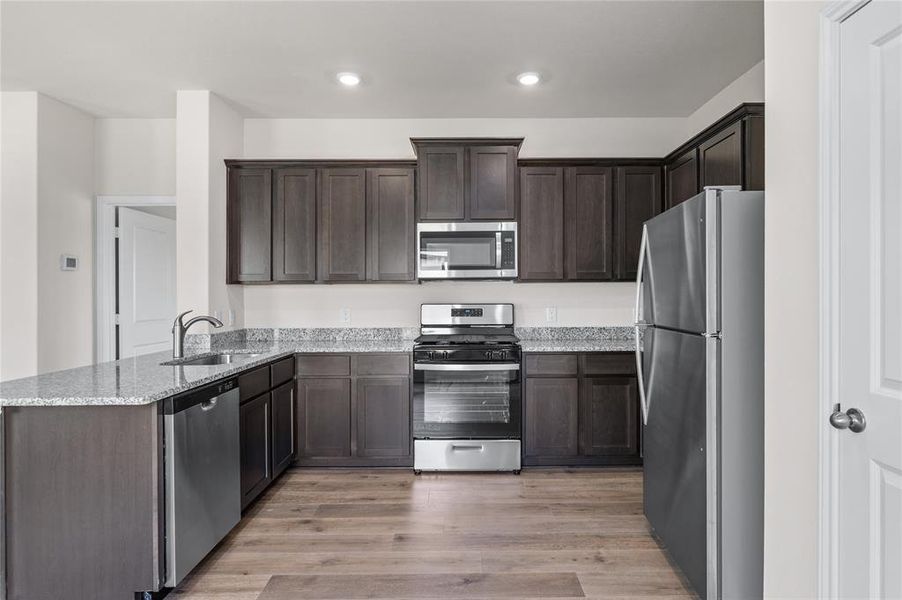 Kitchen featuring appliances with stainless steel finishes, light wood-style floors, light stone countertops, dark brown cabinetry, and a peninsula