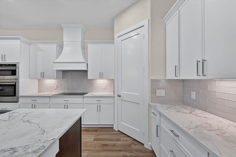 Kitchen featuring white cabinetry, light-toned countertops with distinctive veining, a decorative range hood, and stainless steel built-in appliances Kitchen featuring white cabinetry, light-toned countertops with distinctive veining, a decorative range hood, and stainless steel built-in appliances