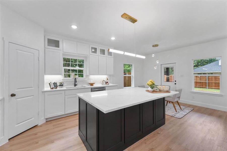 Modern kitchen featuring a large central island with a solid surface countertop, white cabinetry with upper glass-front accents, wood-finish flooring, and recessed lighting Modern kitchen featuring a large central island with a solid surface countertop, white cabinetry with upper glass-front accents, wood-finish flooring, and recessed lighting