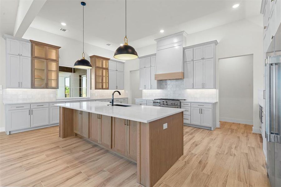 Kitchen featuring glass insert cabinets, a kitchen island with sink, light wood finished floors, light stone counters, and recessed lighting Kitchen featuring glass insert cabinets, a kitchen island with sink, light wood finished floors, light stone counters, and recessed lighting