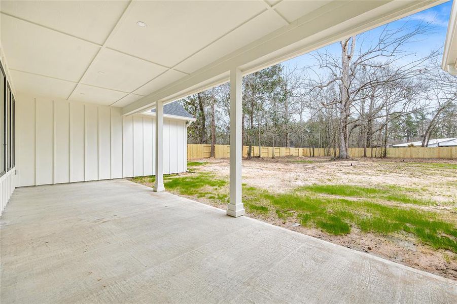 This photo showcases a covered back patio area of a home with white siding and a gray shingled roof. It features a spacious open area with grass and dirt, ideal for outdoor activities or landscaping. The patio is supported by a single column, and multiple windows offer views from inside the house.