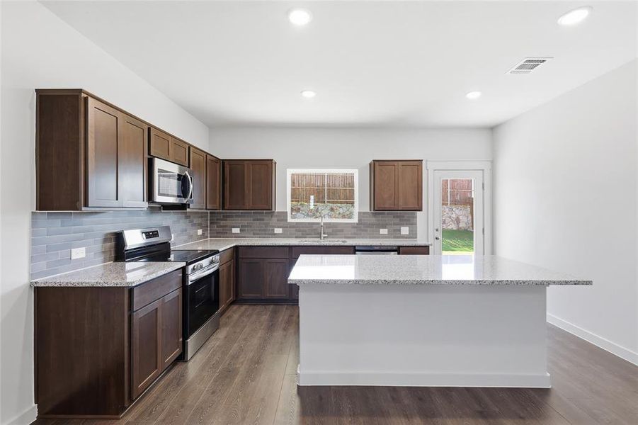 Spacious kitchen featuring dark wood cabinetry, light-colored countertops, stainless steel appliances, a tiled backsplash, and an island with bar seating