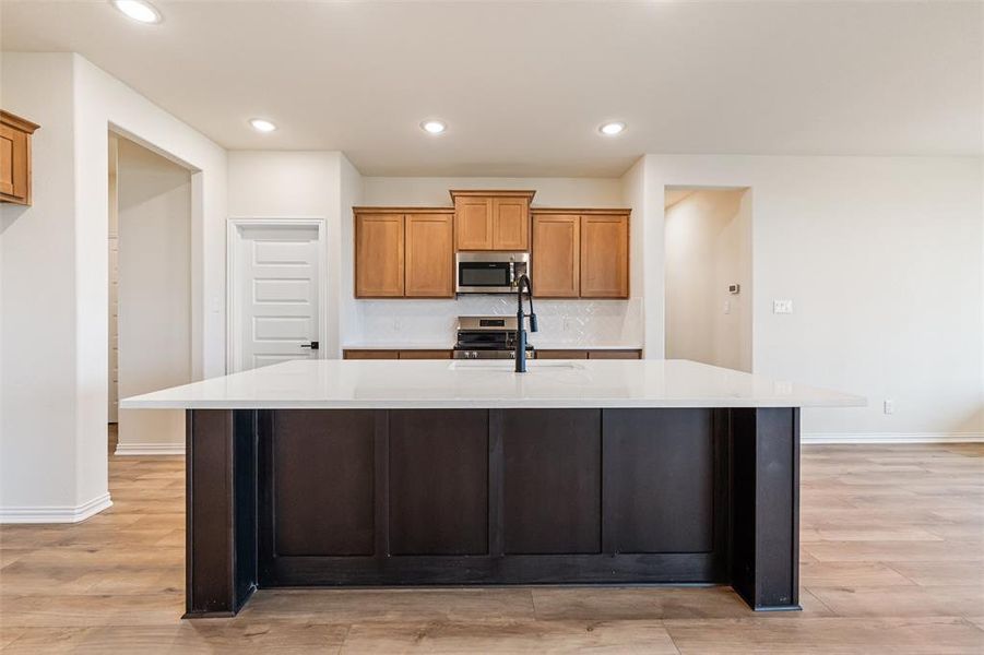 Kitchen featuring brown cabinetry, a center island with sink, light stone countertops, recessed lighting, and appliances with stainless steel finishes