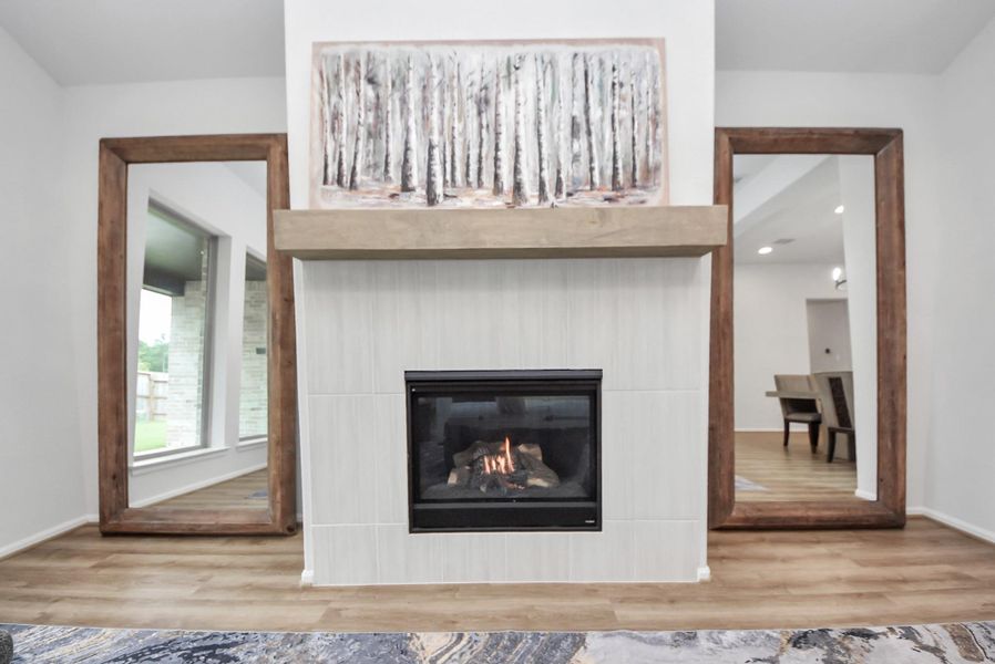 This photo showcases a cozy living space featuring a modern gas fireplace set in a light-colored tile surround. The room is accented with large mirrors framed in wood, enhancing the sense of space. A piece of artwork depicting birch trees adds a touch of nature and elegance above the mantel. The room has a soft, neutral palette with wooden flooring, creating a warm and inviting atmosphere.
