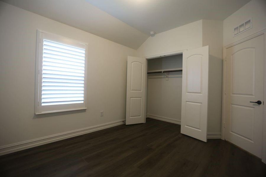 Interior room featuring wood-finish flooring, a window with plantation shutters, vaulted ceilings, a built-in closet with double bifold doors, and white trim