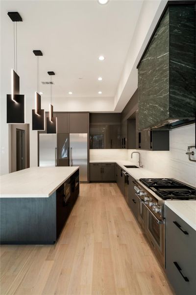 This modern kitchen features sleek black cabinetry, a spacious island with pendant lighting, stainless steel appliances, and elegant hardwood flooring. The design offers ample counter space and a minimalist aesthetic.