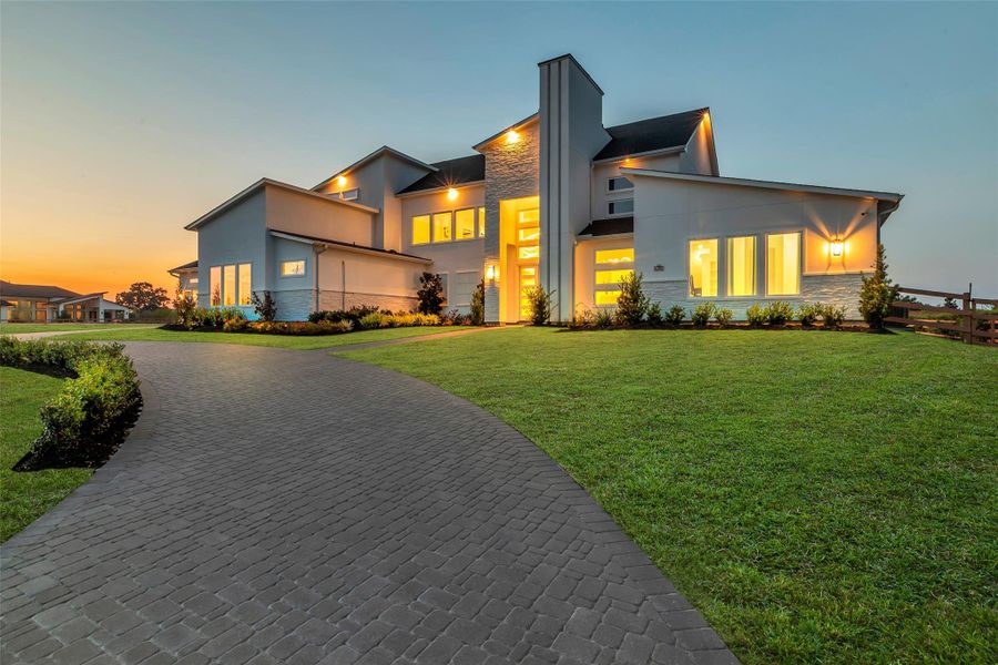 Elegant front elevation with modern architectural lines, stone accents, extended driveway, and illuminated entryway at dusk.