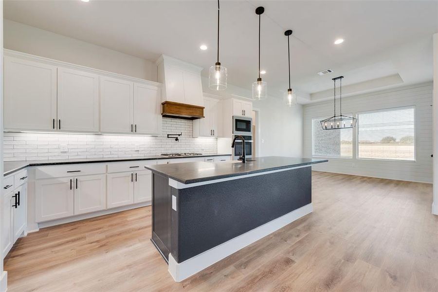 Kitchen featuring white cabinets, a center island with sink, hanging light fixtures, light hardwood / wood-style flooring, and tasteful backsplash