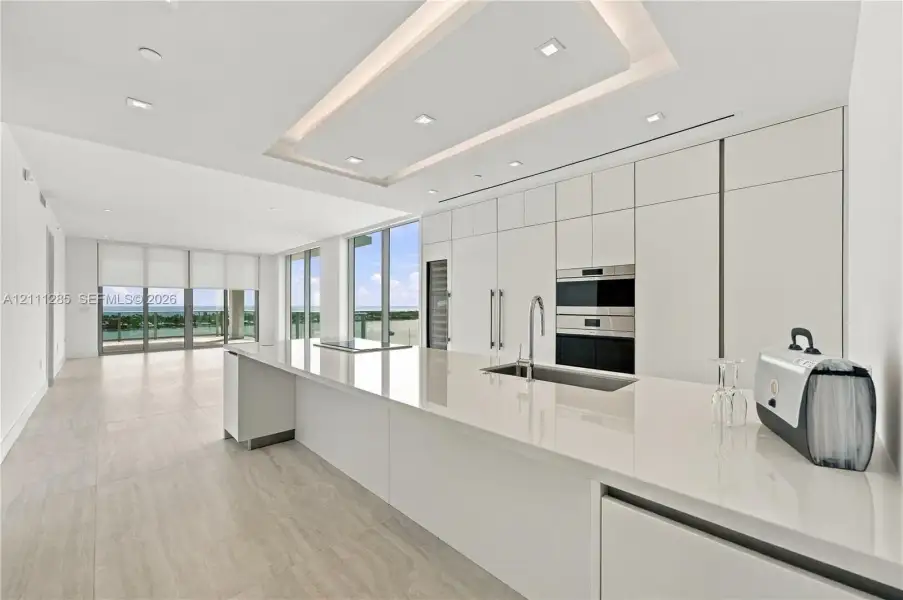 Modern kitchen featuring seamless flat-panel cabinetry, a spacious solid-surface island, built-in stainless steel appliances, recessed tray ceiling lighting, and large-format tile flooring Modern kitchen featuring seamless flat-panel cabinetry, a spacious solid-surface island, built-in stainless steel appliances, recessed tray ceiling lighting, and large-format tile flooring