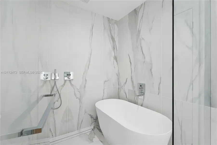 Freestanding white soaking tub with wall-mounted chrome fixtures, floor-to-ceiling large-format stone-finish tile surround, and built-in linear floor drain