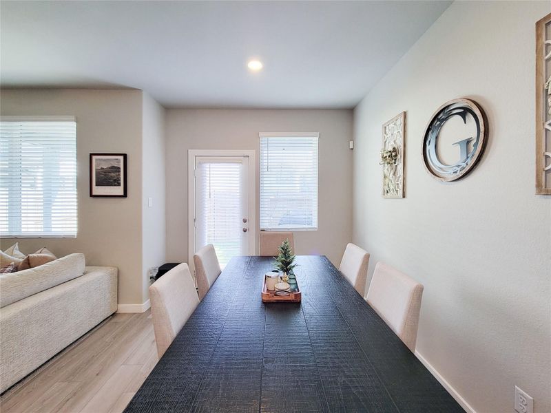 Bright dining room features large windows allowing natural light, light wood flooring, and neutral walls adorned with minimalist decor. Adjacent to a cozy living space, perfect for entertaining.