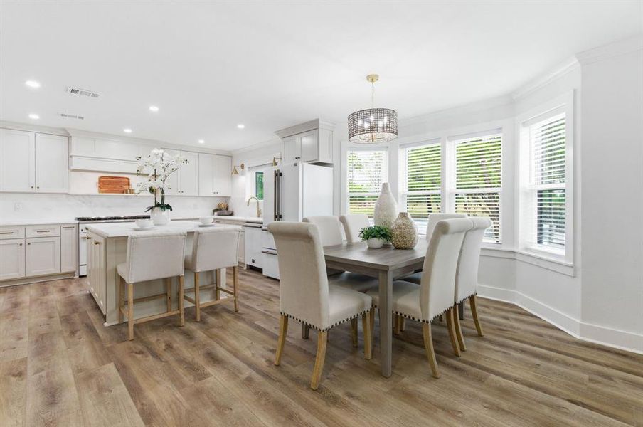 This open-concept area features light wood-style flooring, a kitchen with white cabinetry and an island, and a dining space with a bay window This open-concept area features light wood-style flooring, a kitchen with white cabinetry and an island, and a dining space with a bay window