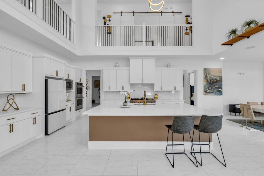 Kitchen & Media Room View: Sleek designer kitchen with oversized island, gold accents, and a view of the upstairs media room framed by statement barn doors.
