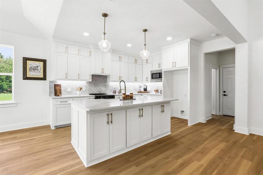 Kitchen featuring an island with a light-toned countertop, white cabinetry with gold-toned hardware, wood-finish flooring, and recessed lighting Kitchen featuring an island with a light-toned countertop, white cabinetry with gold-toned hardware, wood-finish flooring, and recessed lighting