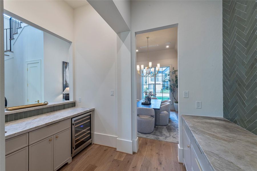 A well-appointed bar  offers extensive cabinetry, stone countertops, a beverage refrigerator, and a striking tiled accent wall, providing both storage and functionality between the kitchen and dining room.
