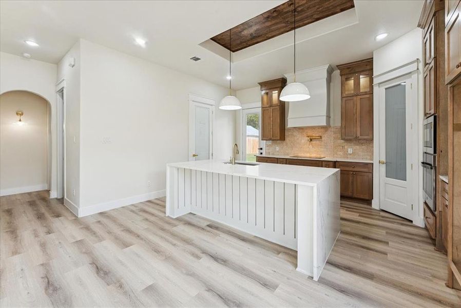 Kitchen featuring glass insert cabinets, brown cabinetry, a kitchen island with sink, arched walkways, and light stone countertops