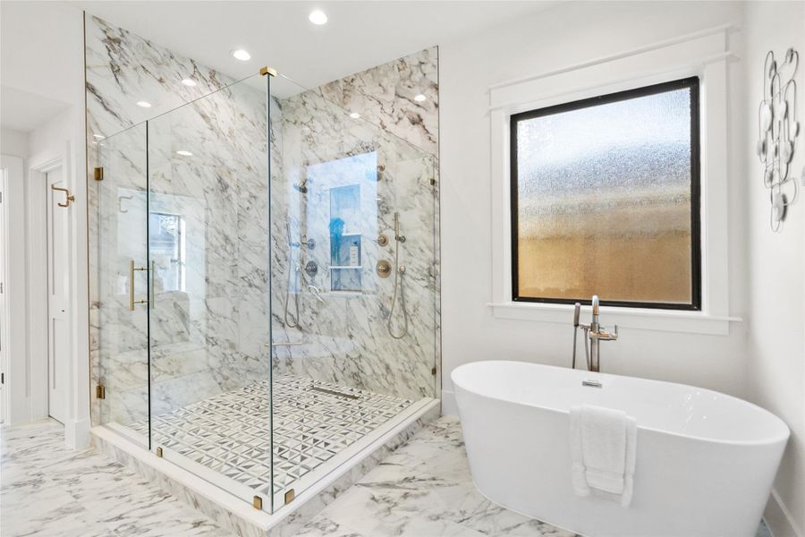 This primary bath is your personal sanctuary—an oversized glass-enclosed shower with statement marble tile sits alongside a sculptural freestanding soaking tub beneath a frosted window. Patterned flooring, refined fixtures, and abundant natural light create a calm, high-end space designed for unwinding in style.