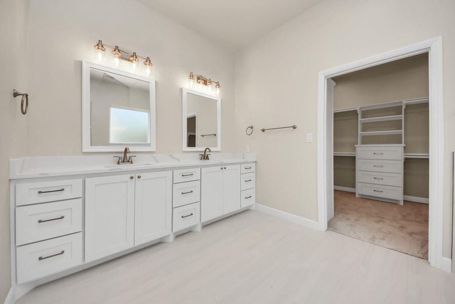This primary bathroom features a double vanity with modern fixtures, dual mirrors, and ample storage. It includes an adjacent walk-in closet with built-in shelves and drawers, offering plenty of organization space.