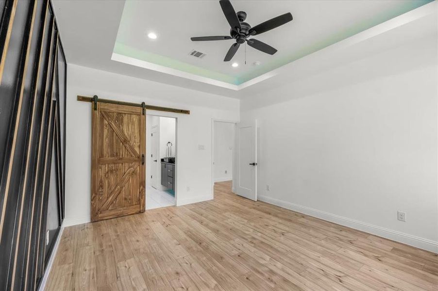 Unfurnished bedroom with light wood-style floors, a ceiling fan, recessed lighting, a barn door, and a raised ceiling