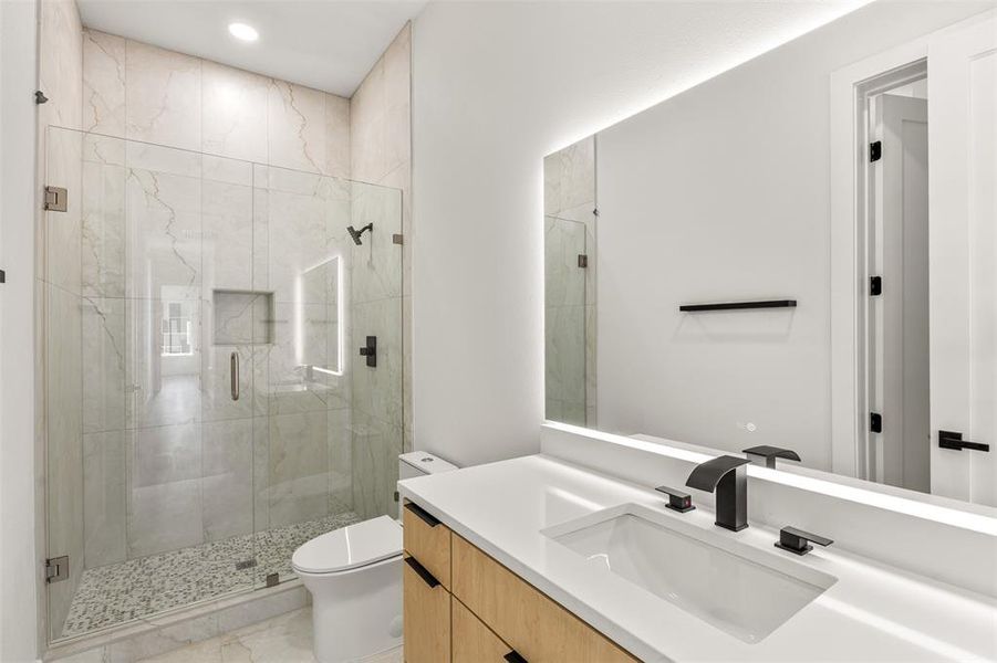 Modern bathroom featuring a frameless glass shower enclosure, large format tile walls, pebble-finish shower floor, an illuminated mirror, and a wood-finish vanity with a white countertop and matte black fixtures