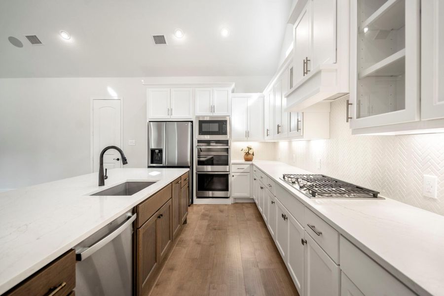 This modern kitchen features dual-tone cabinetry, stainless steel appliances, and a spacious island. It offers ample counter space, double built-in ovens, a gas cooktop, and under cabinet lighting.
