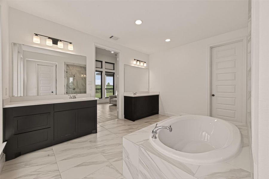 This spacious bathroom features elegant marble floors, a luxurious soaking tub, dual vanities with modern lighting, and a large walk-in shower. The design is bright and contemporary, offering a relaxing atmosphere.
