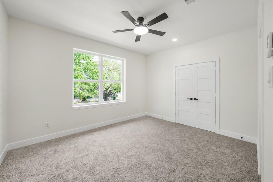 This photo shows a bright, spacious room with neutral walls and carpet flooring. It features a large window with a view of greenery, a modern ceiling fan, and double doors, offering a clean and inviting space.