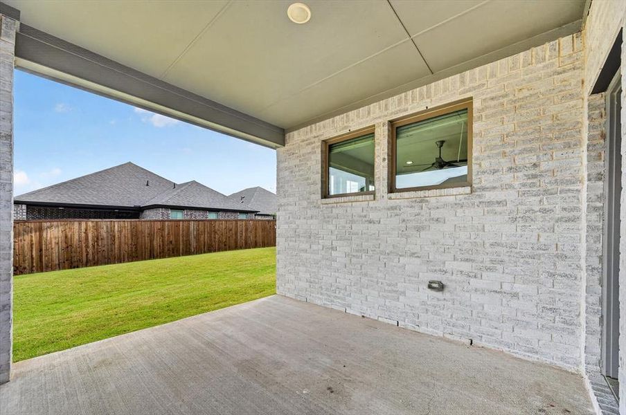 Covered patio with a concrete surface, white brick exterior, two windows with dark frames, and a manicured lawn enclosed by a wooden fence