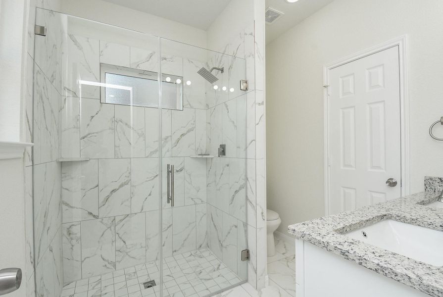 Modern bathroom with marble tiles, glass shower enclosure, granite countertop, white cabinets, and a toilet behind a closed door. Modern bathroom with marble tiles, glass shower enclosure, granite countertop, white cabinets, and a toilet behind a closed door.