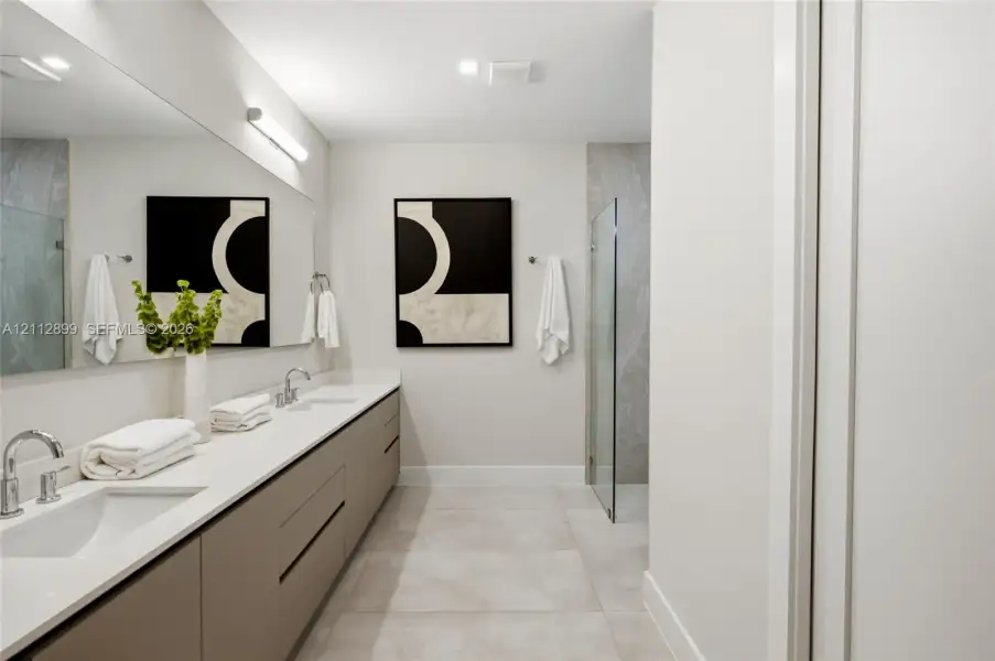Dual-sink floating vanity with neutral cabinetry, expansive wall-mounted mirror, light-toned stone-finish flooring, and glass-enclosed shower with full-height stone-finish wall cladding