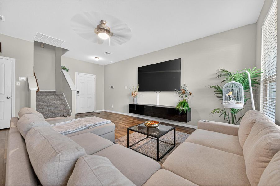 This cozy living room features a large sectional sofa, modern decor, and a wall-mounted TV. It has hardwood floors, a ceiling fan, and ample natural light from the windows. A staircase leads to the upper floor, adding to the spacious feel.