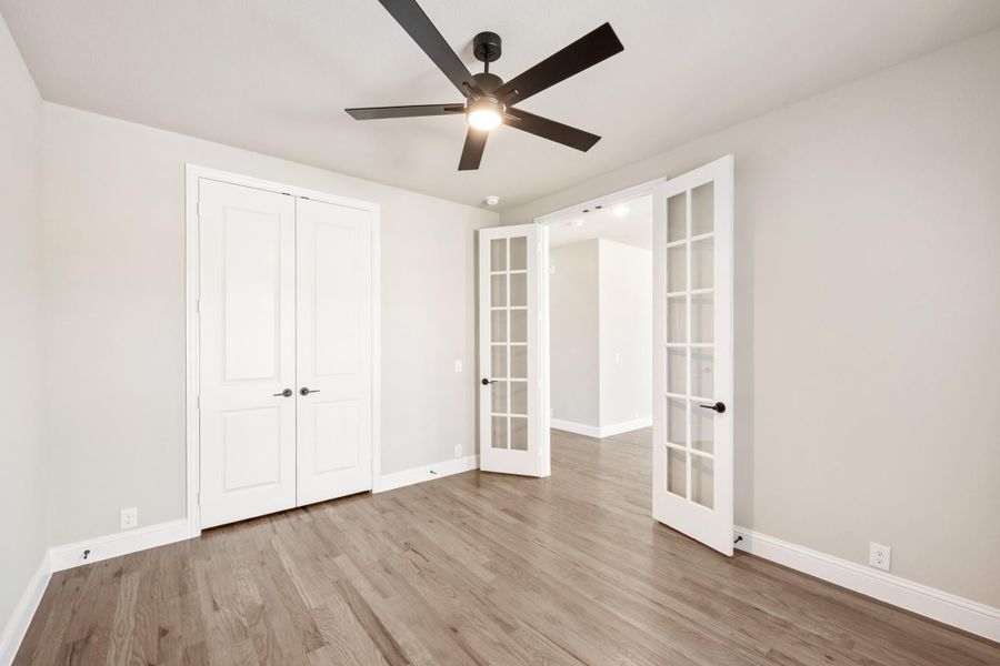 Empty bedroom with hardwood floors, ceiling fan, double closet doors, and open French doors leading to hallway