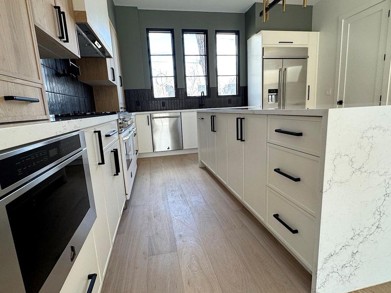 Kitchen featuring light stone countertops, appliances with stainless steel finishes, wall chimney range hood, light wood-type flooring, and backsplash