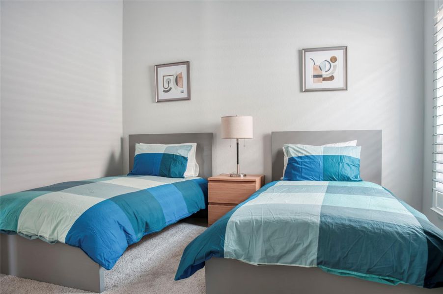 Bright and cozy bedroom with two twin beds, featuring modern blue and teal bedding. A shared nightstand with a lamp sits between the beds, and there's soft carpet underfoot. Natural light streams through a window with shutters, enhancing the room's inviting atmosphere. Bright and cozy bedroom with two twin beds, featuring modern blue and teal bedding. A shared nightstand with a lamp sits between the beds, and there's soft carpet underfoot. Natural light streams through a window with shutters, enhancing the room's inviting atmosphere.