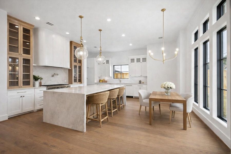 Chef inspired kitchen anchored by a waterfall quartz island, custom white cabinetry, and warm wood accents that add depth and contrast. Glass front upper cabinets, brass pendant lighting, and wide plank hardwood floors create a bright open concept layout with seamless flow to the dining area and expansive black framed windows. *Virtually Staged*