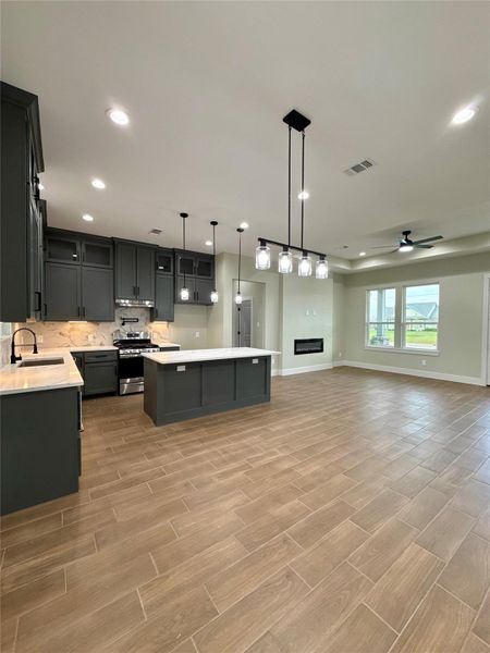 This photo showcases a modern, open-concept kitchen and living area. It features dark cabinetry, a central island, and stylish pendant lighting. The space is complemented by wood-look tile flooring, a modern built-in fireplace, and large windows offering plenty of natural light.