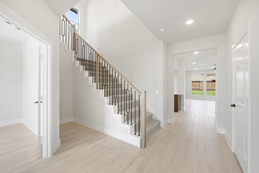 Bright entryway with carpeted staircase, metal balusters, wood railing, and light hardwood floors leading to main living area.