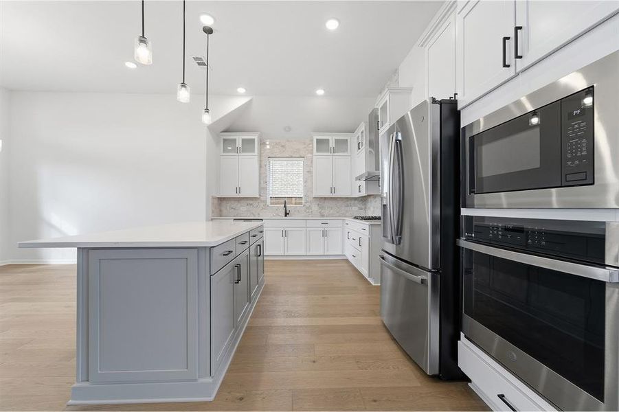 Modern kitchen featuring a light gray island with a white countertop, stainless steel appliances including a double oven and refrigerator, white shaker-style cabinetry, and light wood-finish flooring