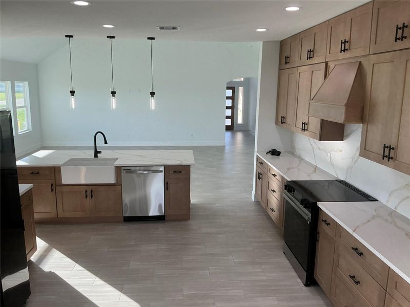This photo shows a modern kitchen with light wood cabinetry, quartz countertops, and a central island with a farmhouse sink. It features stainless steel appliances, pendant lighting, and an open layout leading to a bright living area.
