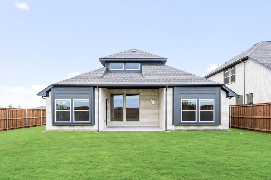 Rear exterior of single-story brick home with gray roof, covered patio, large windows, and fenced backyard lawn