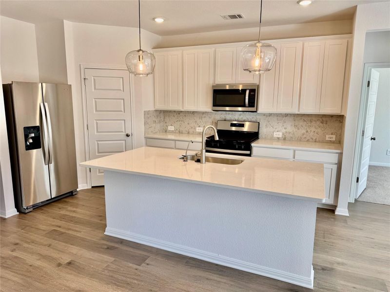 Kitchen with stainless steel appliances, hanging light fixtures, light stone counters, white cabinetry, and an island with sink