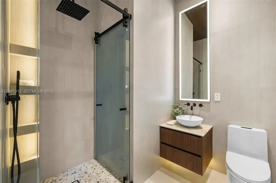 Floating wood-finish vanity with a stone countertop and white vessel sink, large-format tile wall cladding, walk-in shower with matte black hardware and sliding glass door, pebble-stone shower floor, and back-lit rectangular mirror Floating wood-finish vanity with a stone countertop and white vessel sink, large-format tile wall cladding, walk-in shower with matte black hardware and sliding glass door, pebble-stone shower floor, and back-lit rectangular mirror