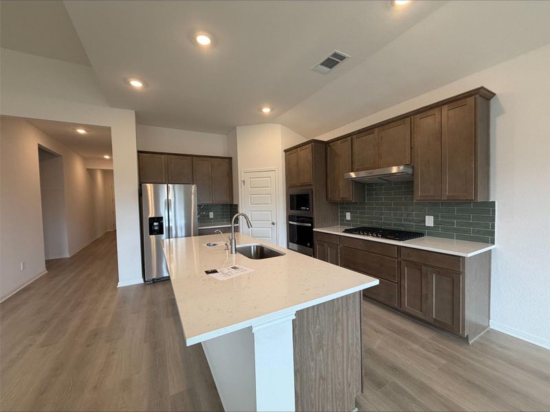 Kitchen featuring decorative backsplash, stainless steel appliances, light stone countertops, an island with sink, and light wood-style flooring