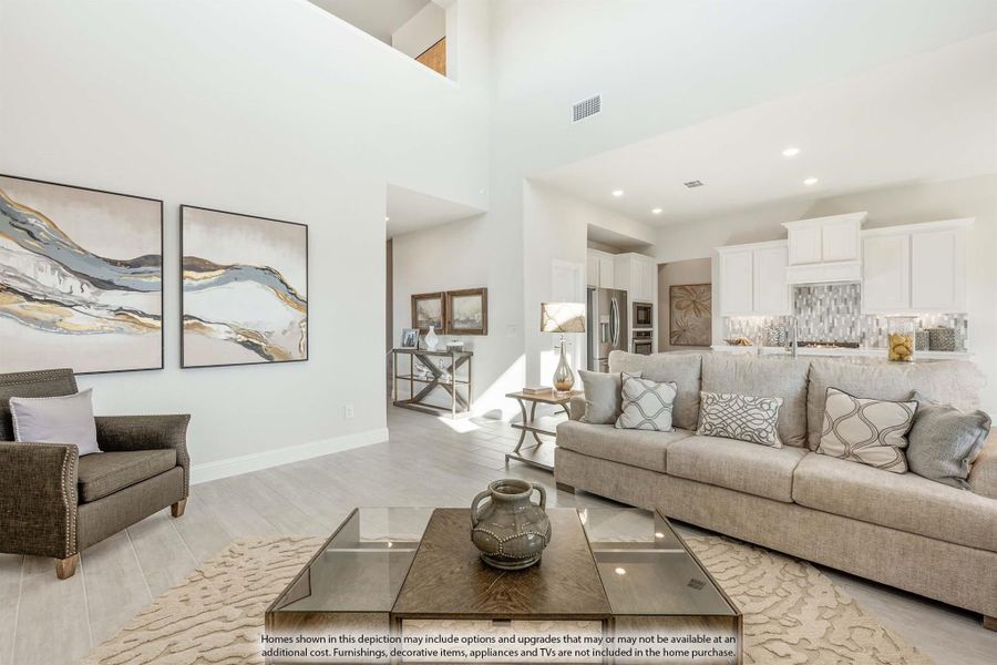 Open-concept living room with high ceilings, neutral sofa, glass coffee table, and white kitchen visible in background