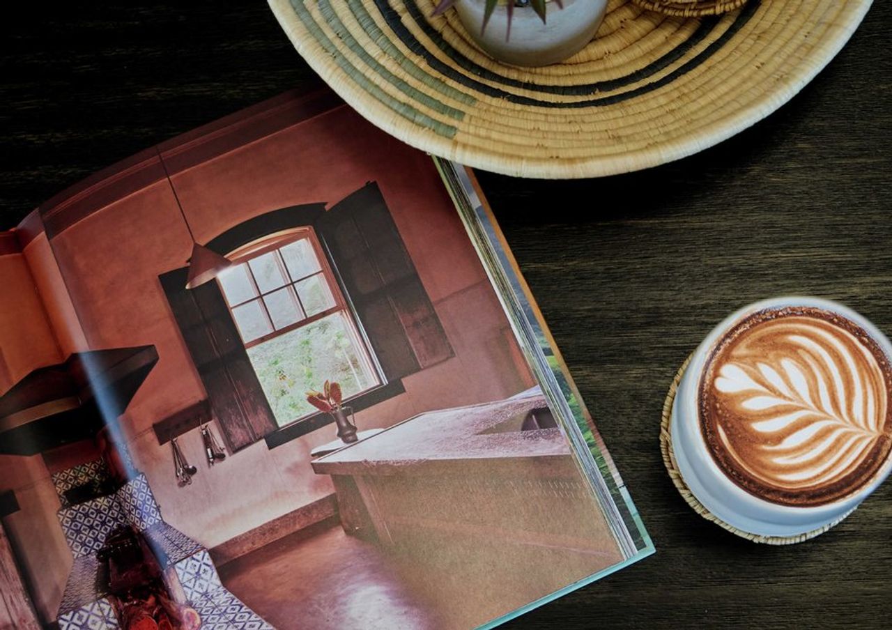 A wooden coffee table with a home decor book opened and a fresh latte in a mug next to it. A wooden coffee table with a home decor book opened and a fresh latte in a mug next to it.