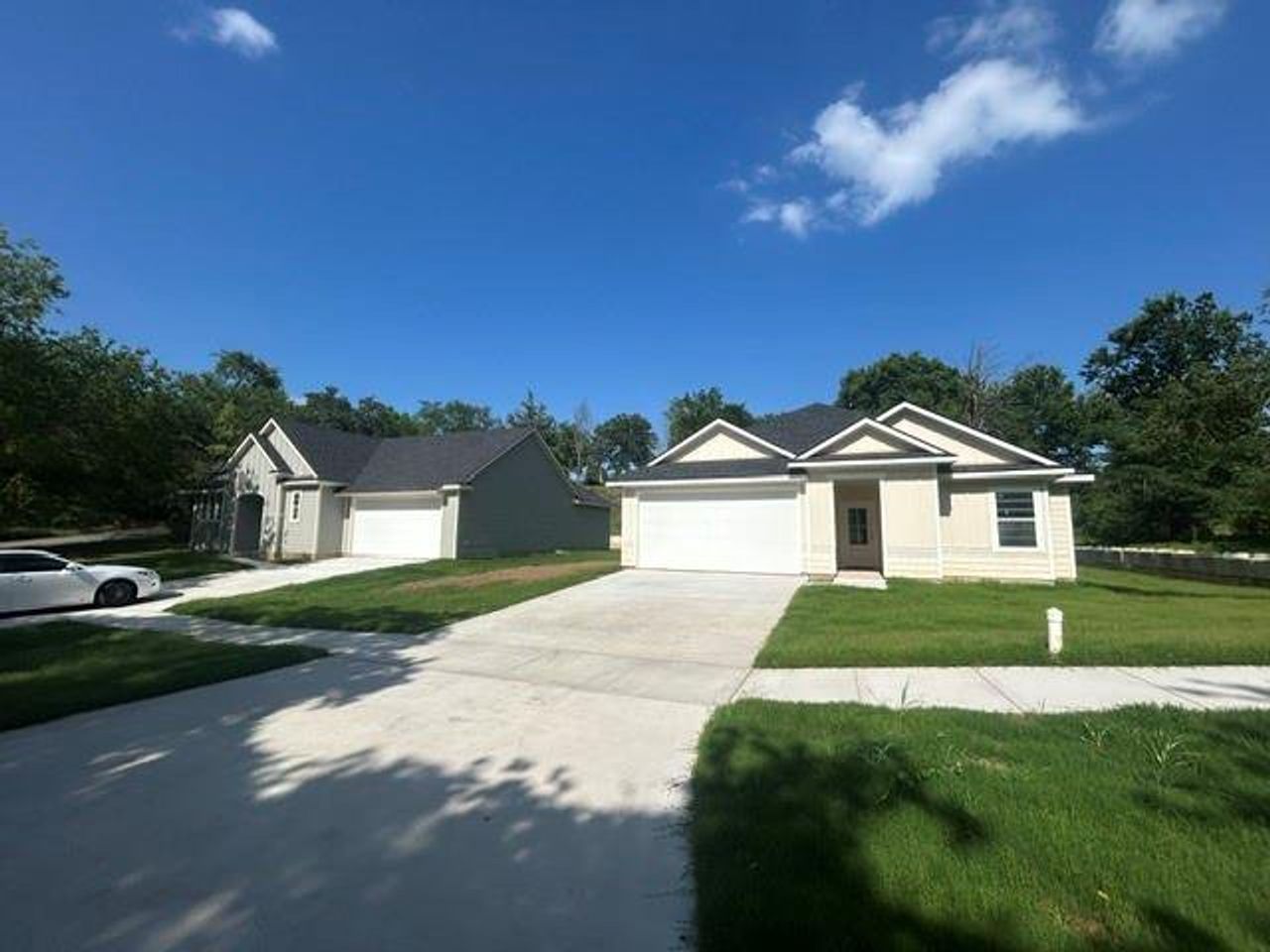 Contemporary residences featuring tan and gray siding, dark shingle roofing, attached two-car garages with white doors, concrete driveways, and green lawns